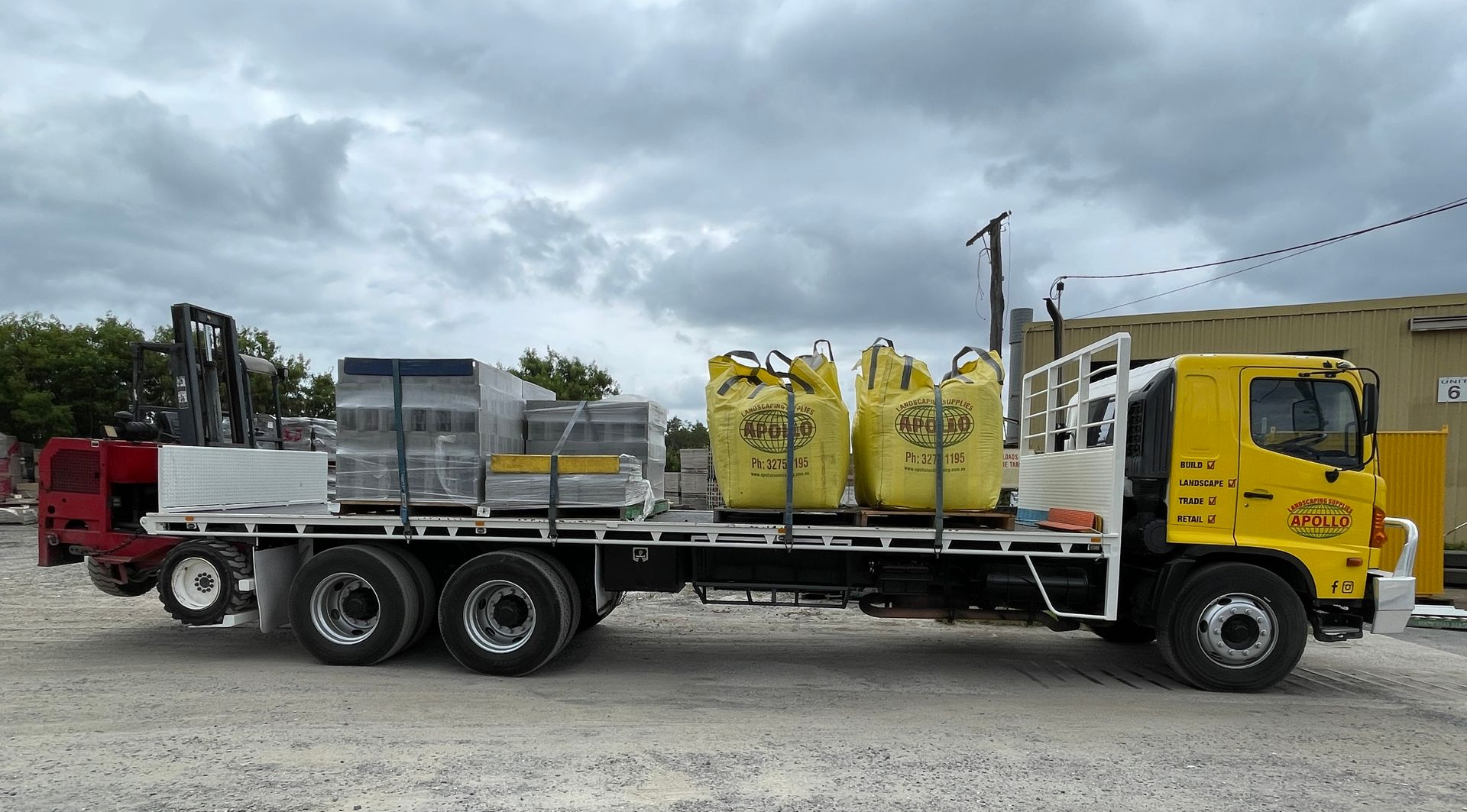 Yellow truck with cargo of bricks and large yellow bags parked outdoors under cloudy sky.