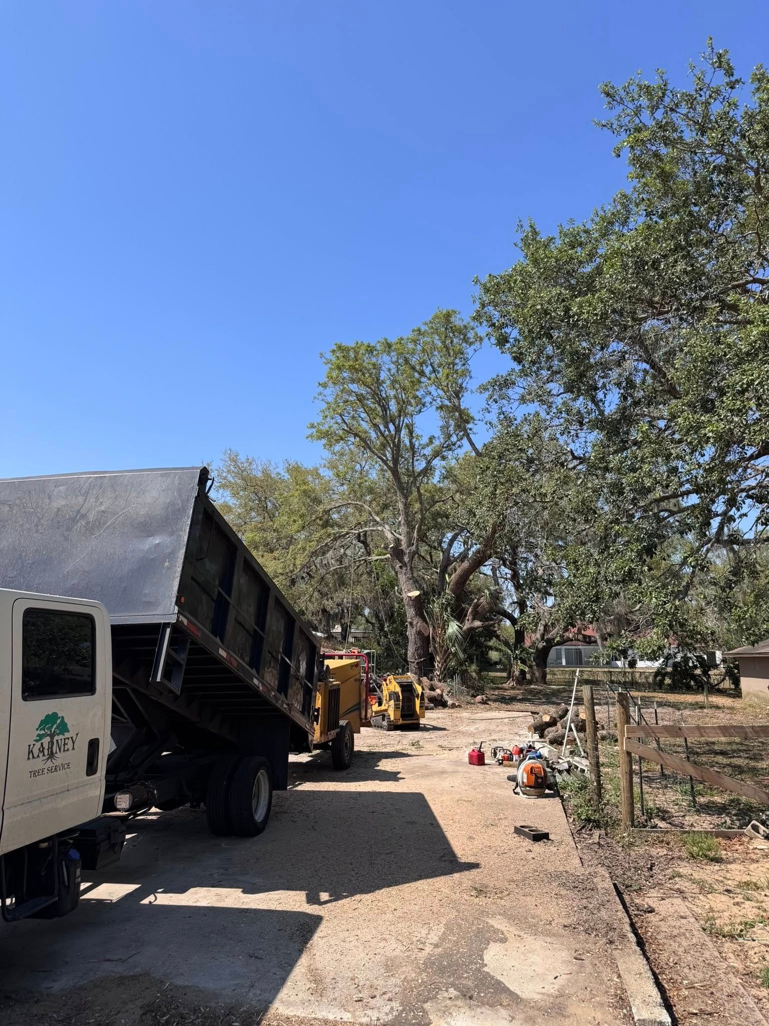 Truck and machinery on a gravel path near trees, with a blue sky overhead.