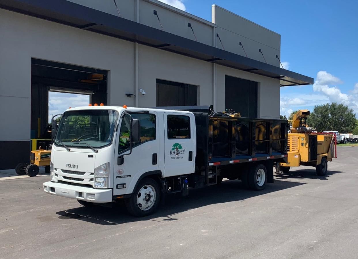 White Isuzu truck with logo parked outside a building, towing a wood chipper on a sunny day.