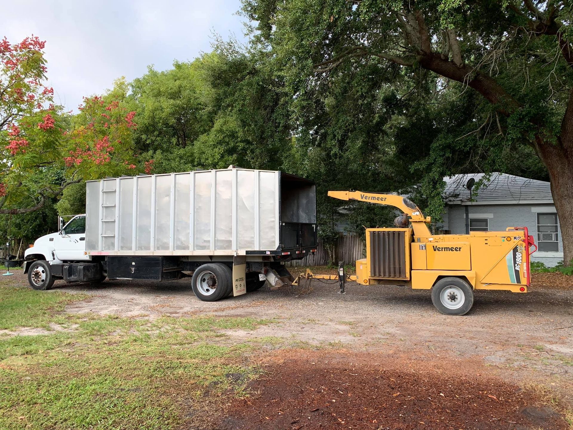 Yellow wood chipper next to a truck with an open metal container, parked on grass near trees.