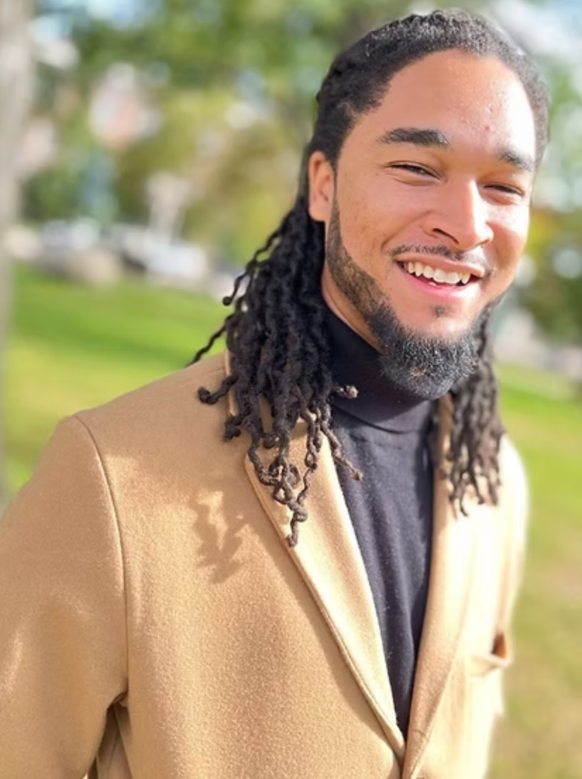 A man with dreadlocks is wearing a tan jacket and a black turtleneck.