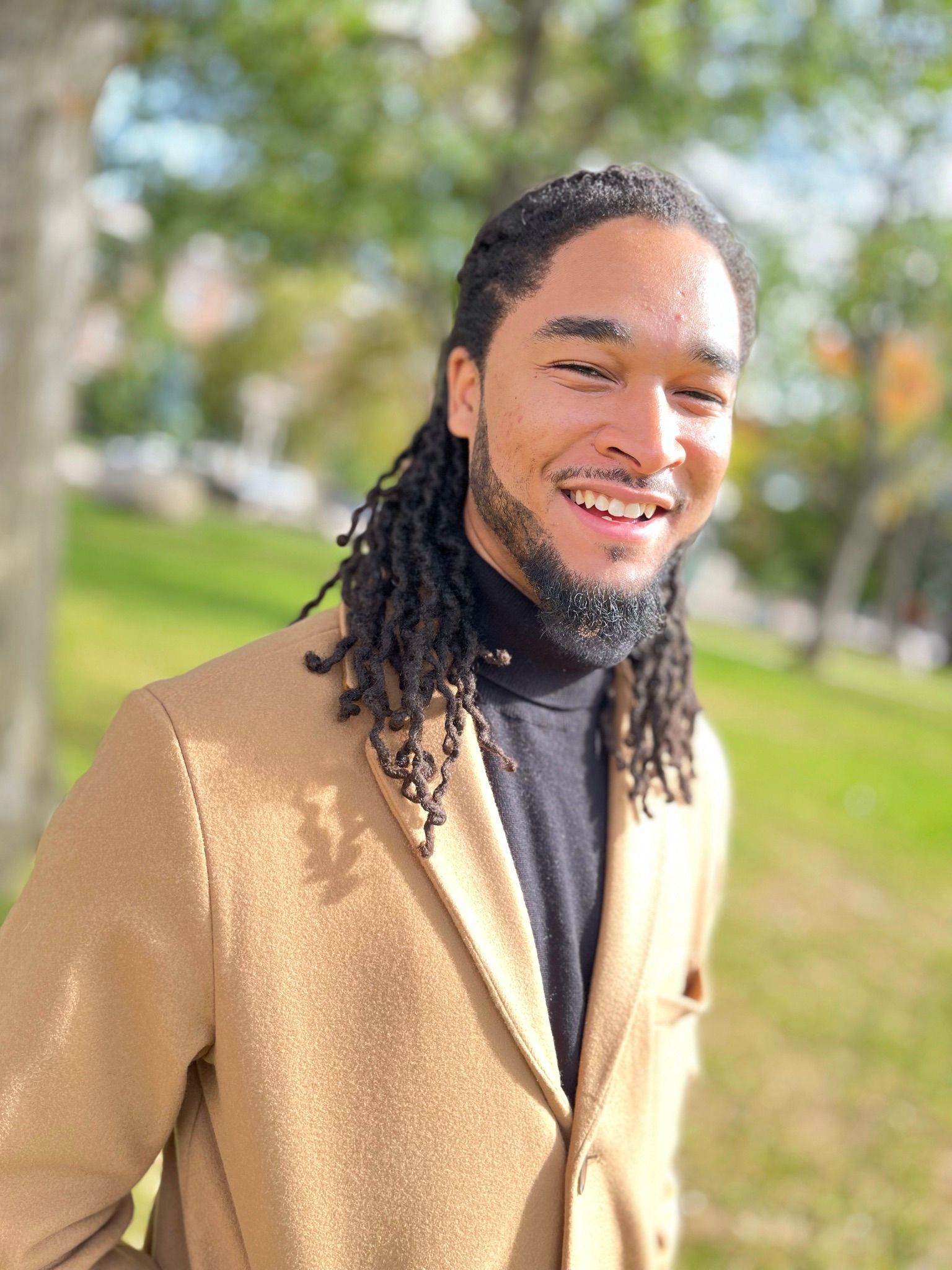 A man with dreadlocks is wearing a tan jacket and a black turtleneck.