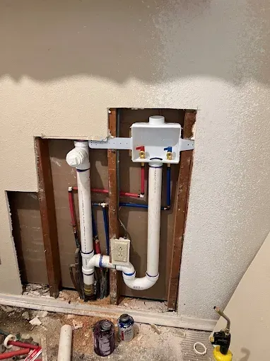 Open plumbing for a washing machine in a wall. White and red pipes, water hookups, and drain.