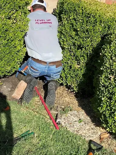 Plumber kneeling by bushes, working on a pipe in a yard. Red and green tools visible.