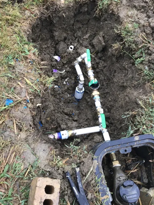 Open irrigation system components in a muddy hole outdoors. Green, white, and bronze pipes, and a black box.