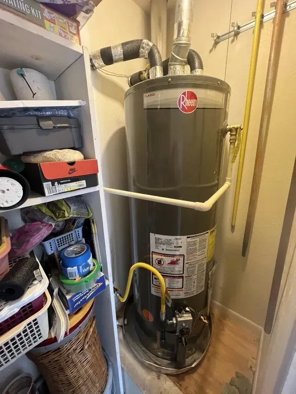 A Rheem water heater in a utility closet with exhaust pipe and yellow gas line. Shelves on the left.