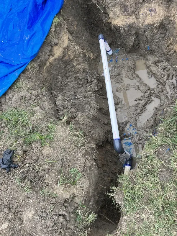 Trench dug in the ground with white PVC pipe, black connectors, and a blue tarp.