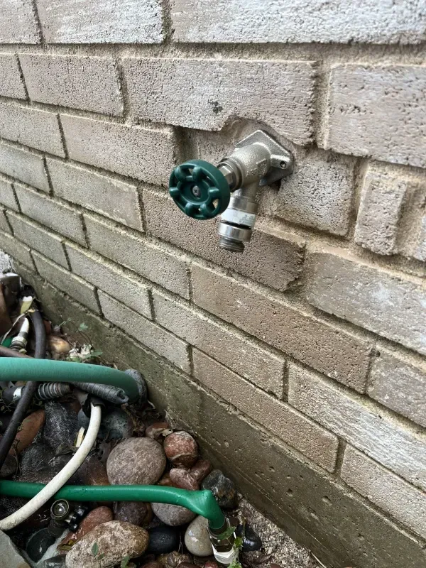 Outdoor spigot on brick wall, green handle, hose attached, set among rocks and garden debris.