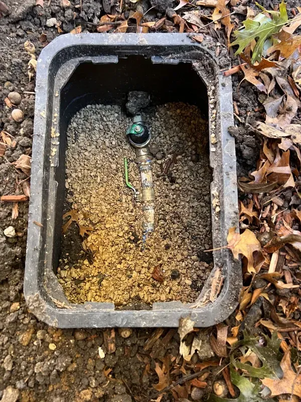 Open irrigation valve box in the ground, filled with gravel, containing valve and pipe, outdoors.