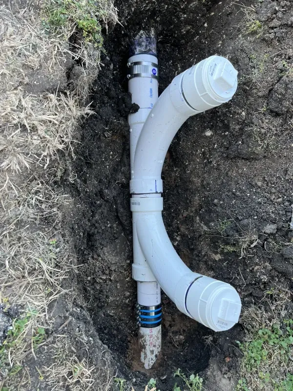 White PVC plumbing pipes installed in a trench in the ground, with curves and connections visible.