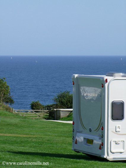 A white caravan is parked in a grassy field campsite with the ocean in the background.