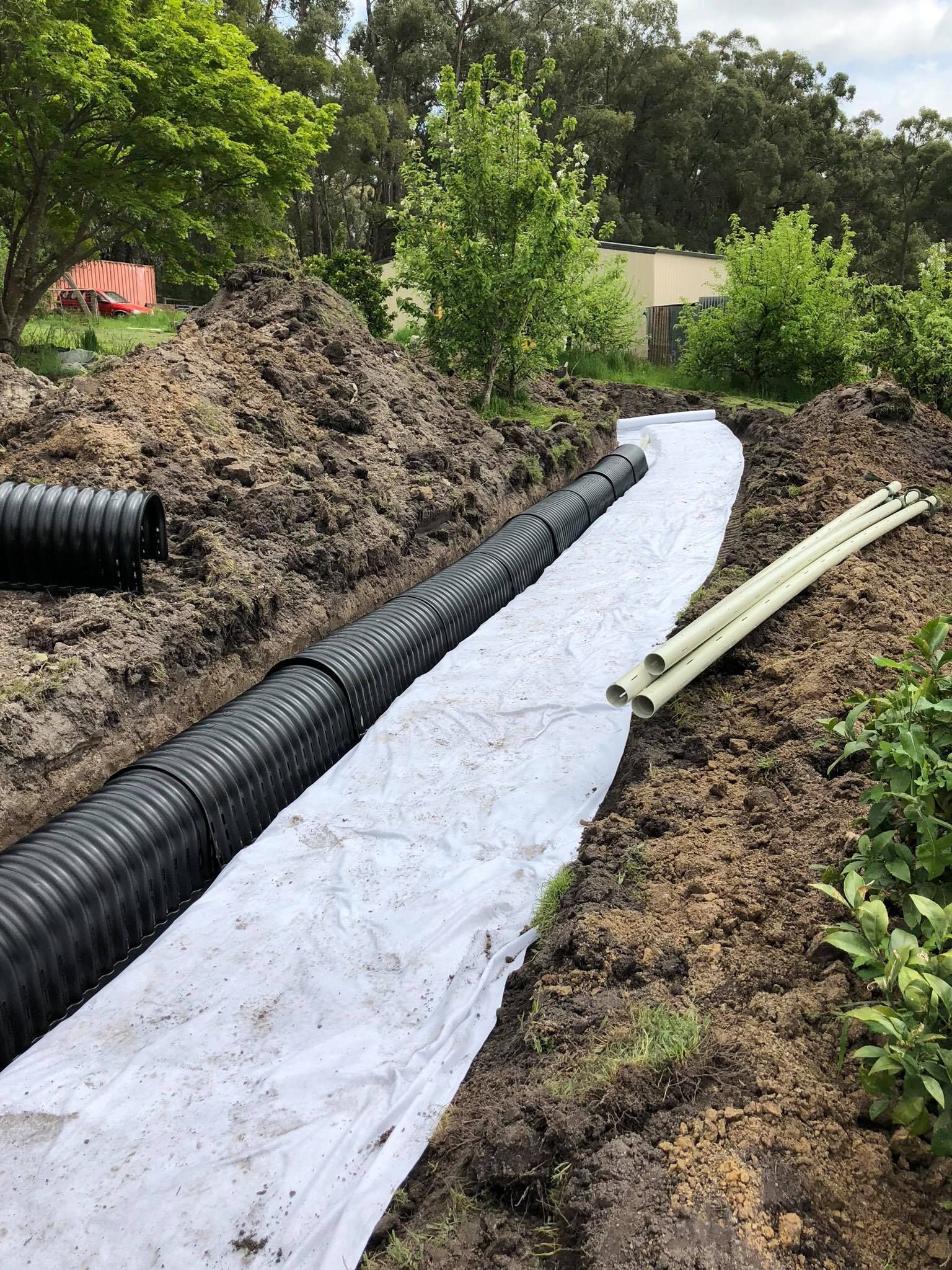Ditch with black drainage pipe, white fabric, and yellow pipes. Dirt surrounds, trees in background.