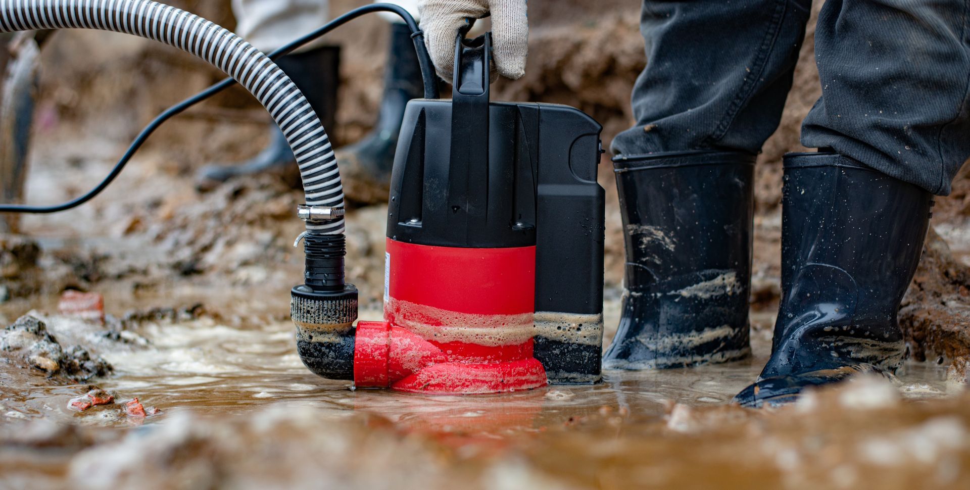 Person in boots using a red and black submersible pump to remove water from a muddy area.