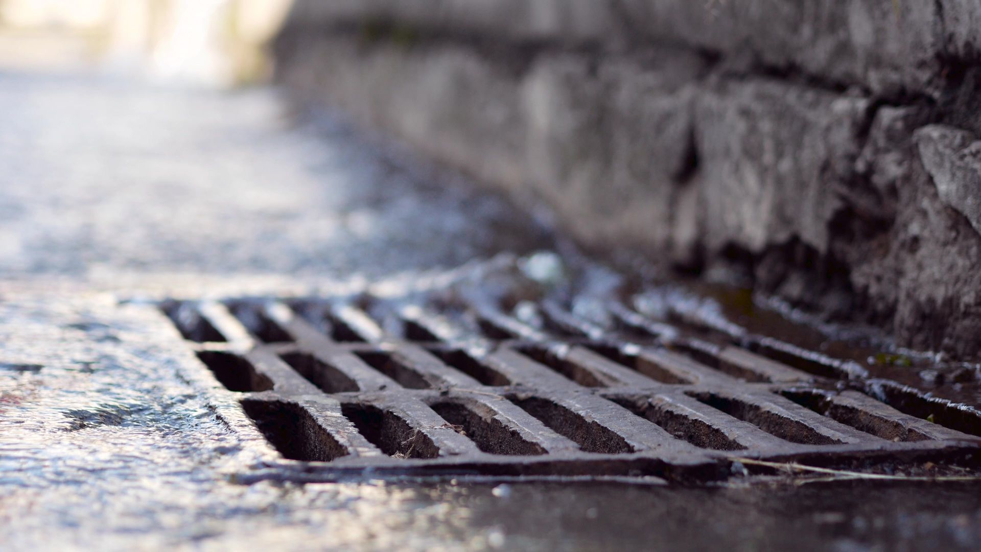 Metal grate drain with water runoff on a concrete surface next to a stone wall.