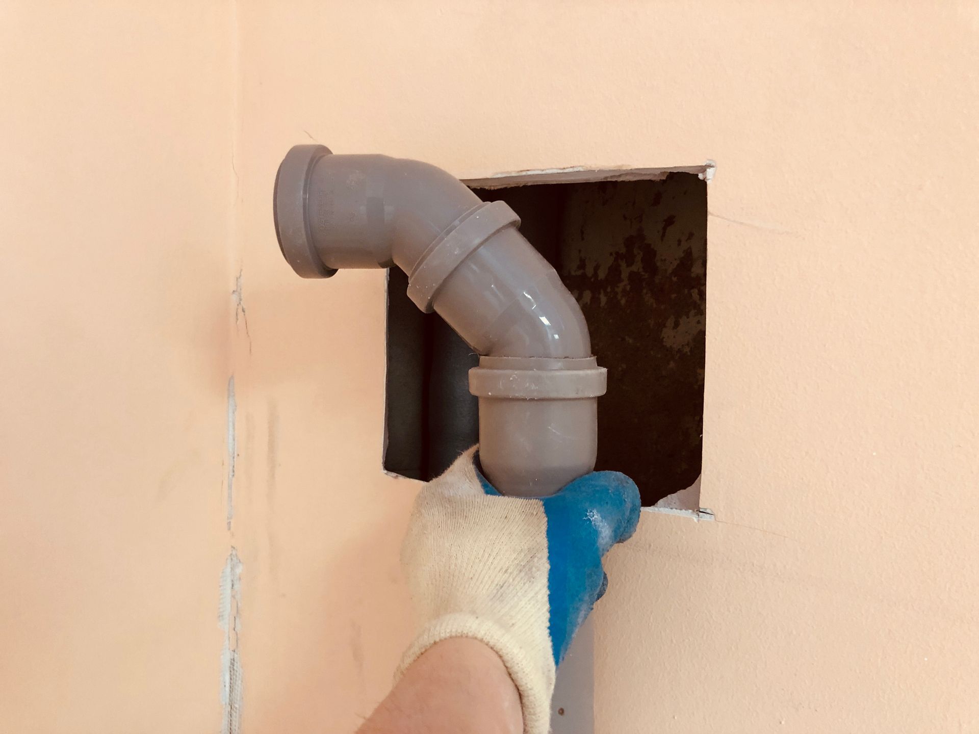 A person in a glove holds a grey pipe, fitting it into a wall opening.