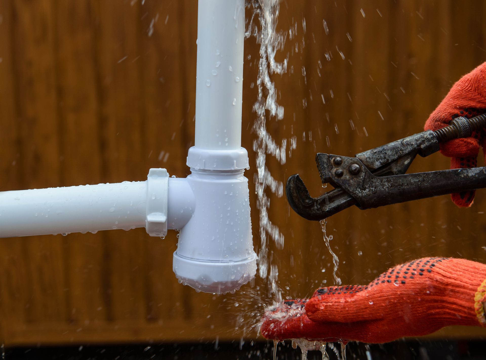 Plumber with red gloves uses wrench on a leaking white pipe, water spraying, brown background.