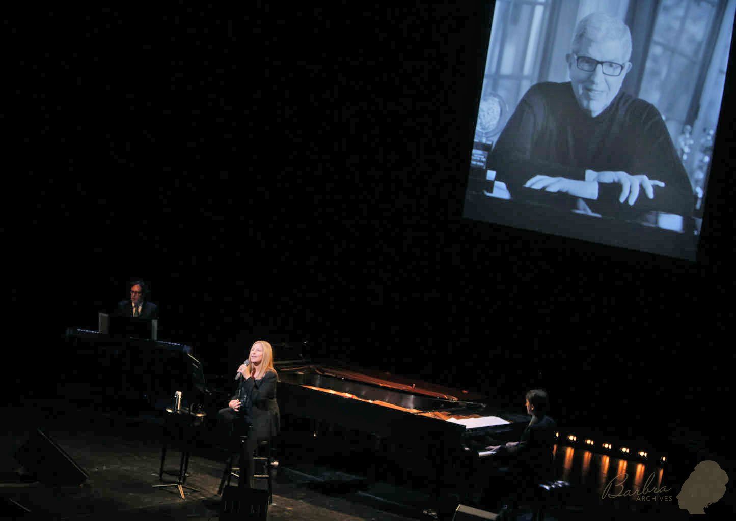 Streisand on stage at Juilliard with Marvin Hamlisch's photograph behind her.
