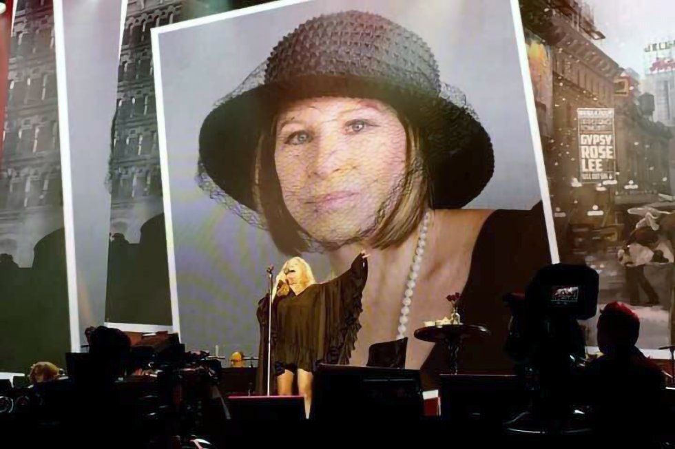 Streisand on stage in London with costume and makeup test for Gypsy projected behind her.