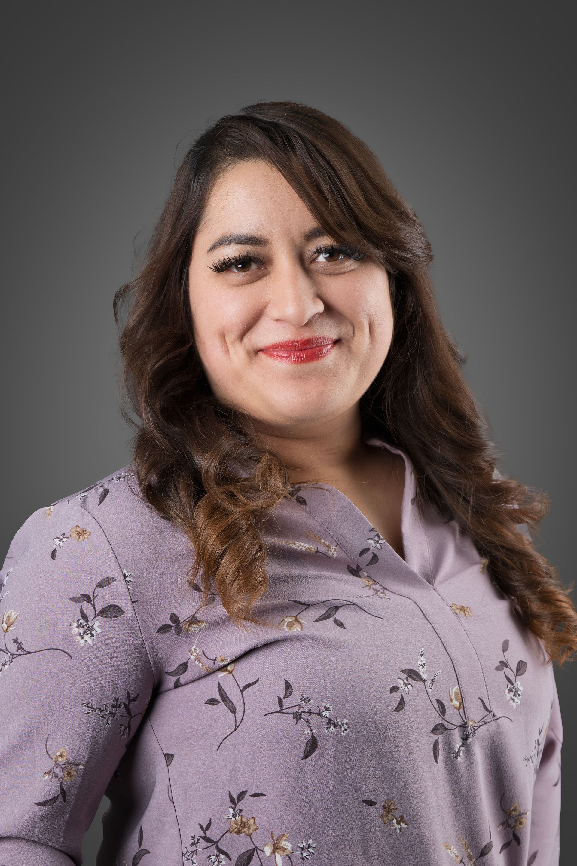 Woman with long brown hair, wearing a purple floral blouse, smiling at the camera.