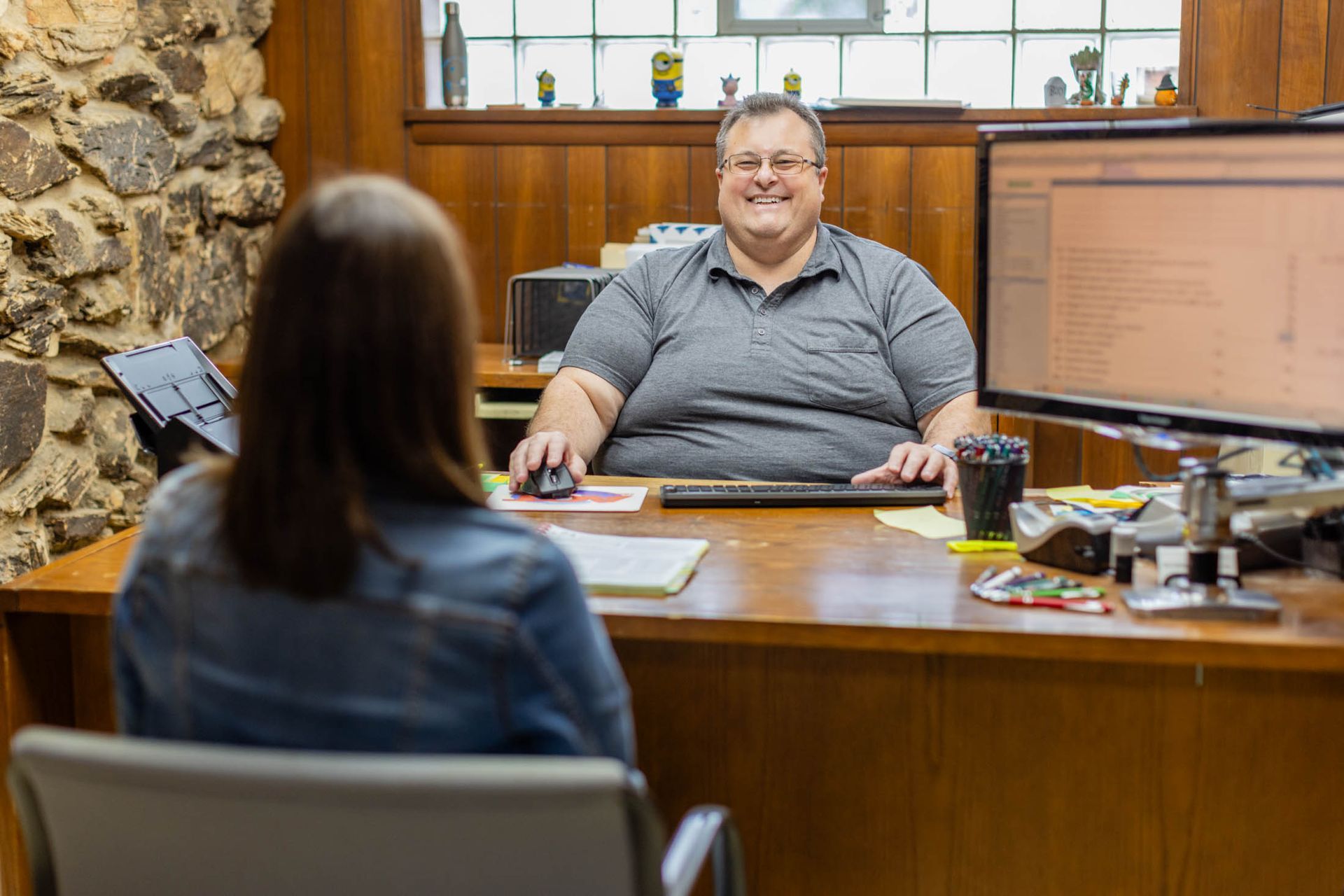 Man in gray shirt smiling, sitting at desk, talking to woman in blue jacket. Interior office setting.