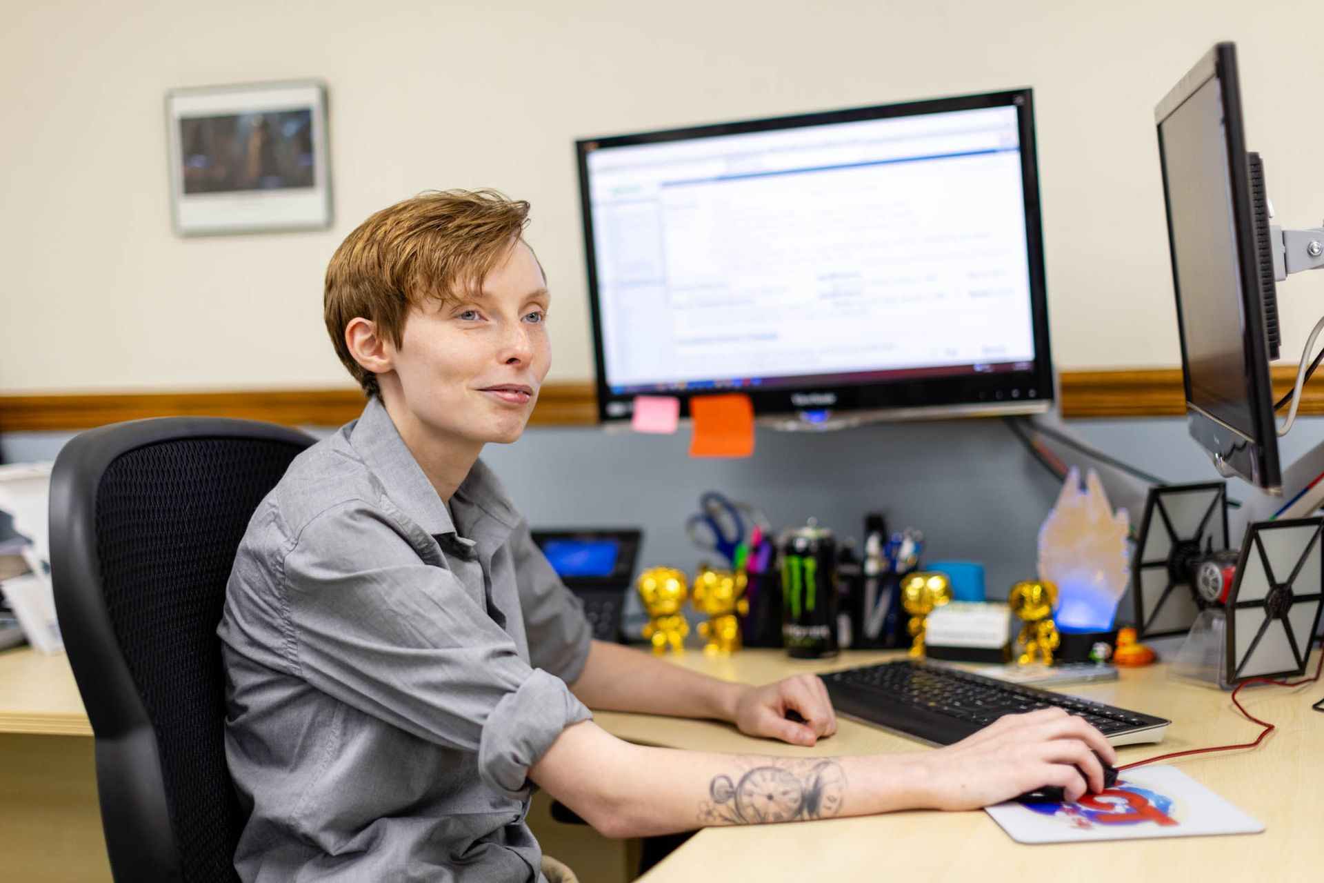 Person with short red hair in office, working at a computer with two monitors. Their left arm has a tattoo.