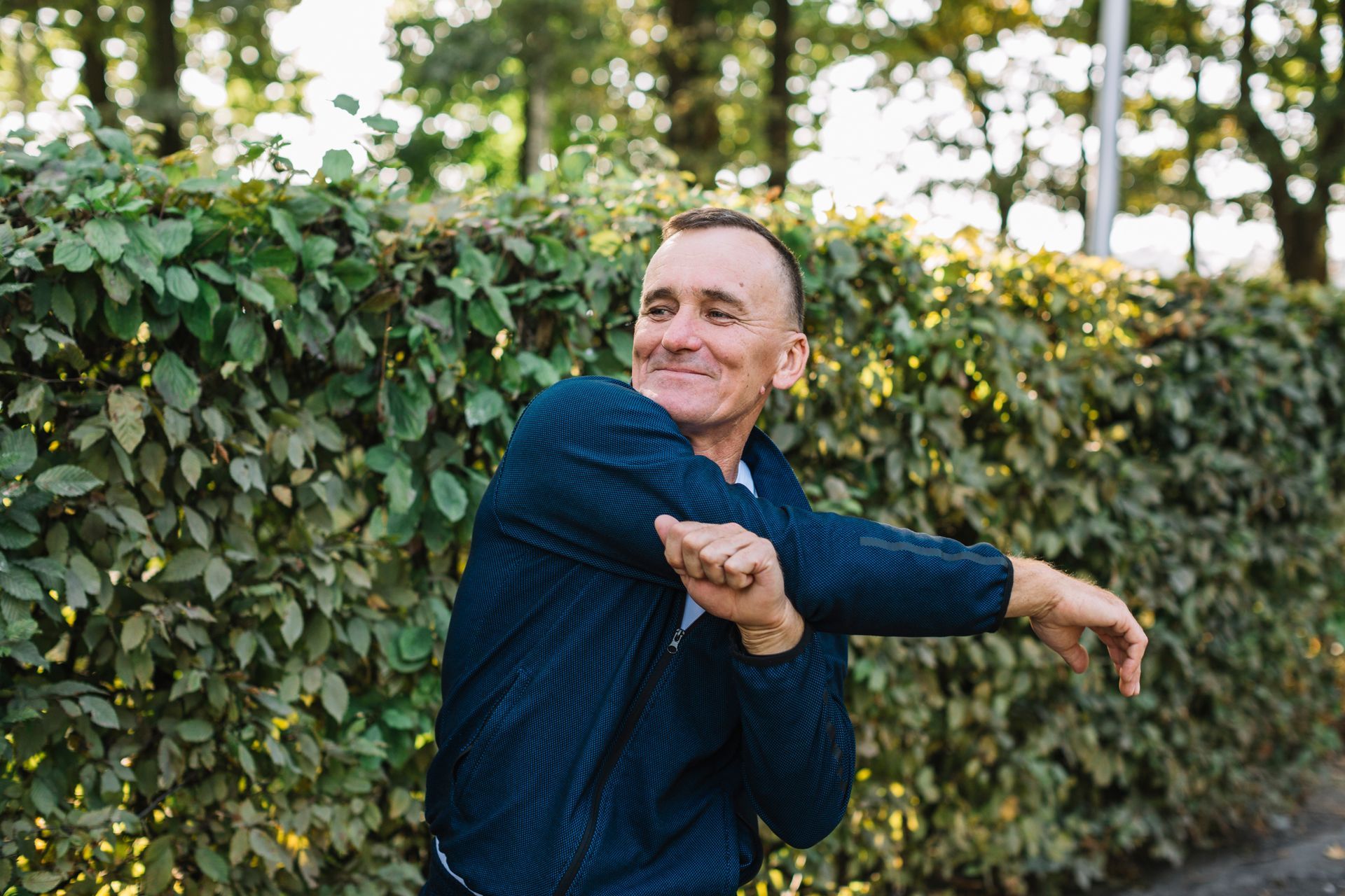 a man in a blue jacket stretches his arms in front of a hedge