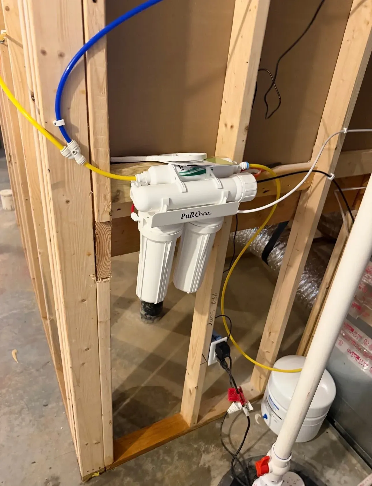 Under-sink water filter system installed between exposed wall studs with blue and yellow hoses.