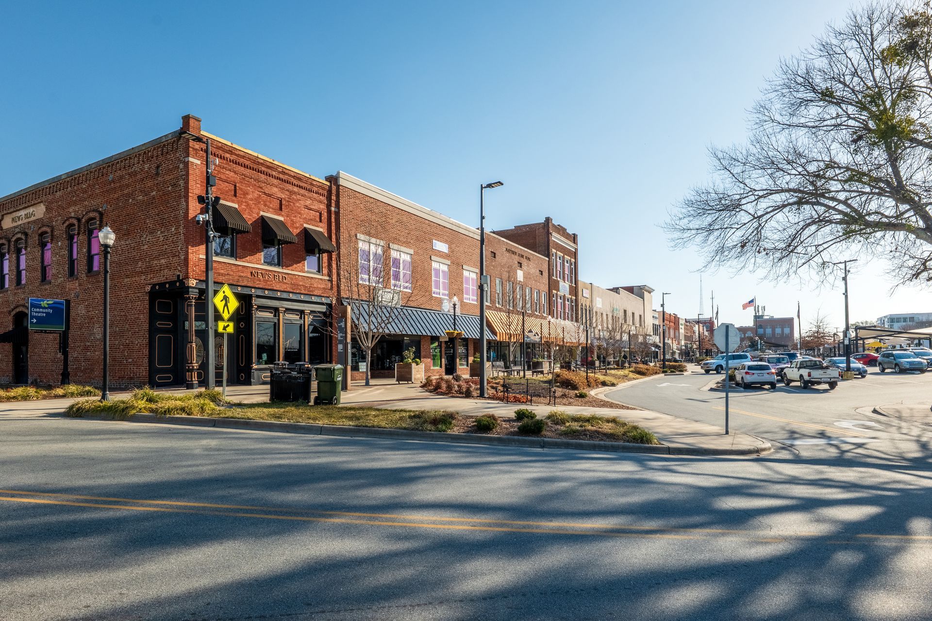 A street-level view of a brick downtown building on a sunny day with parked cars and a large tree on the right.