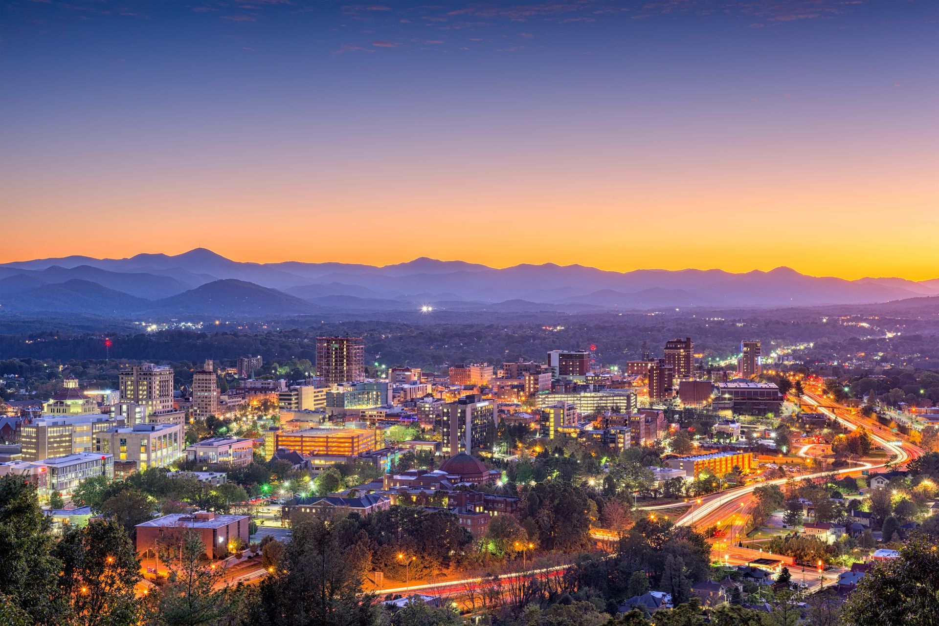 A dusk cityscape of Asheville, North Carolina, illuminated with city lights against a backdrop of mountain silhouettes.