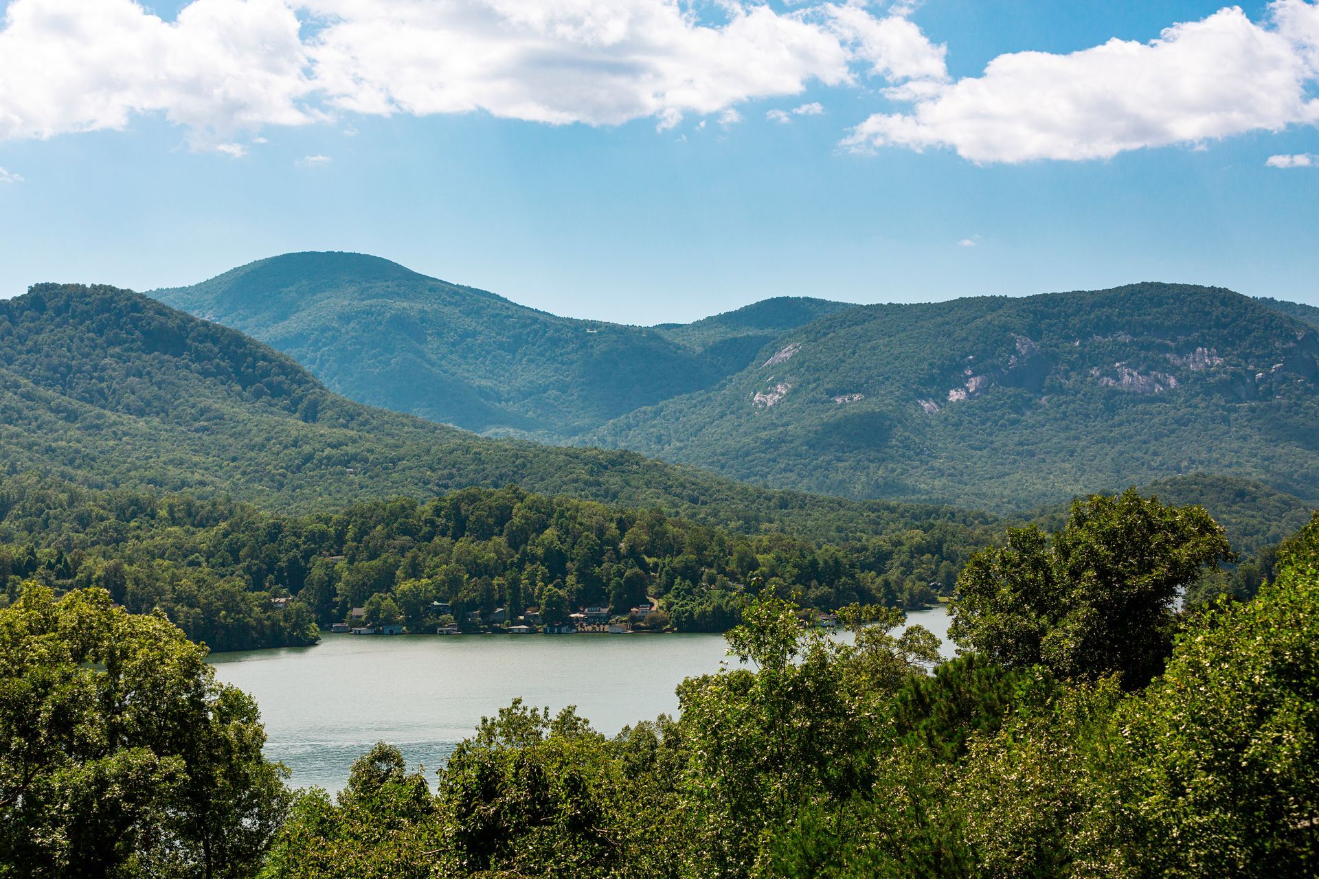 A lush mountain range overlooking a calm lake under a bright, partly cloudy blue sky.