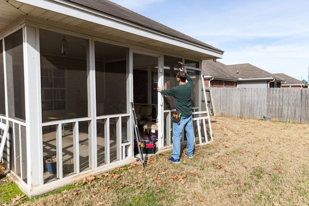 Man installing a screen on a porch. White screens and trim. Brown grass, blue sky.