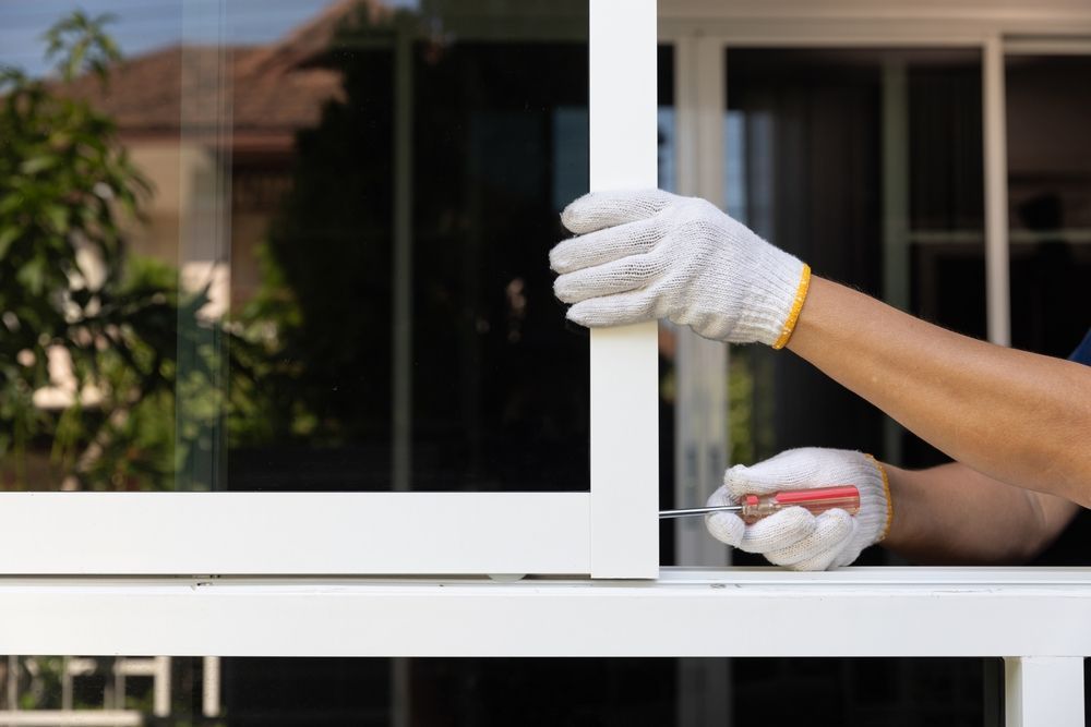 Person in gloves using a screwdriver to adjust a white sliding window frame.