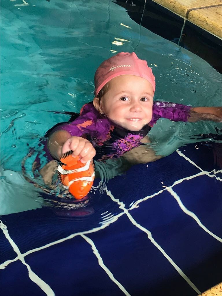 A little girl is swimming in a pool and holding a toy fish.