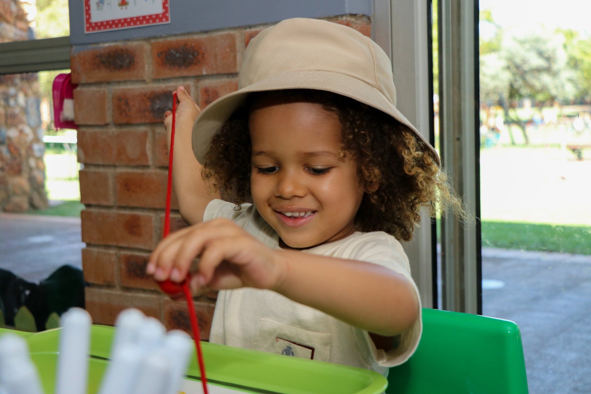 A little girl wearing a hat is playing with a red string.