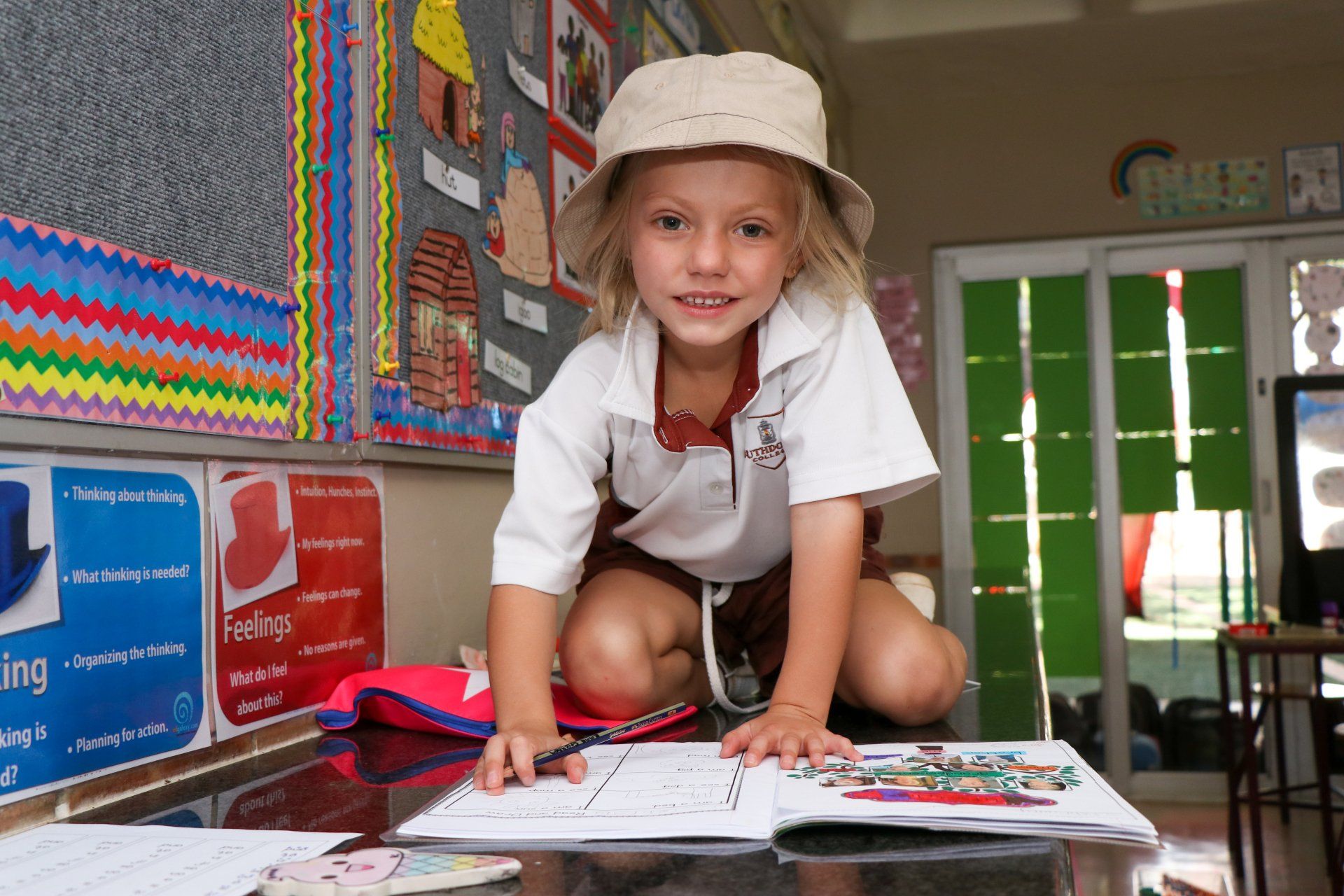A little girl wearing a hat is kneeling on a table in a classroom.