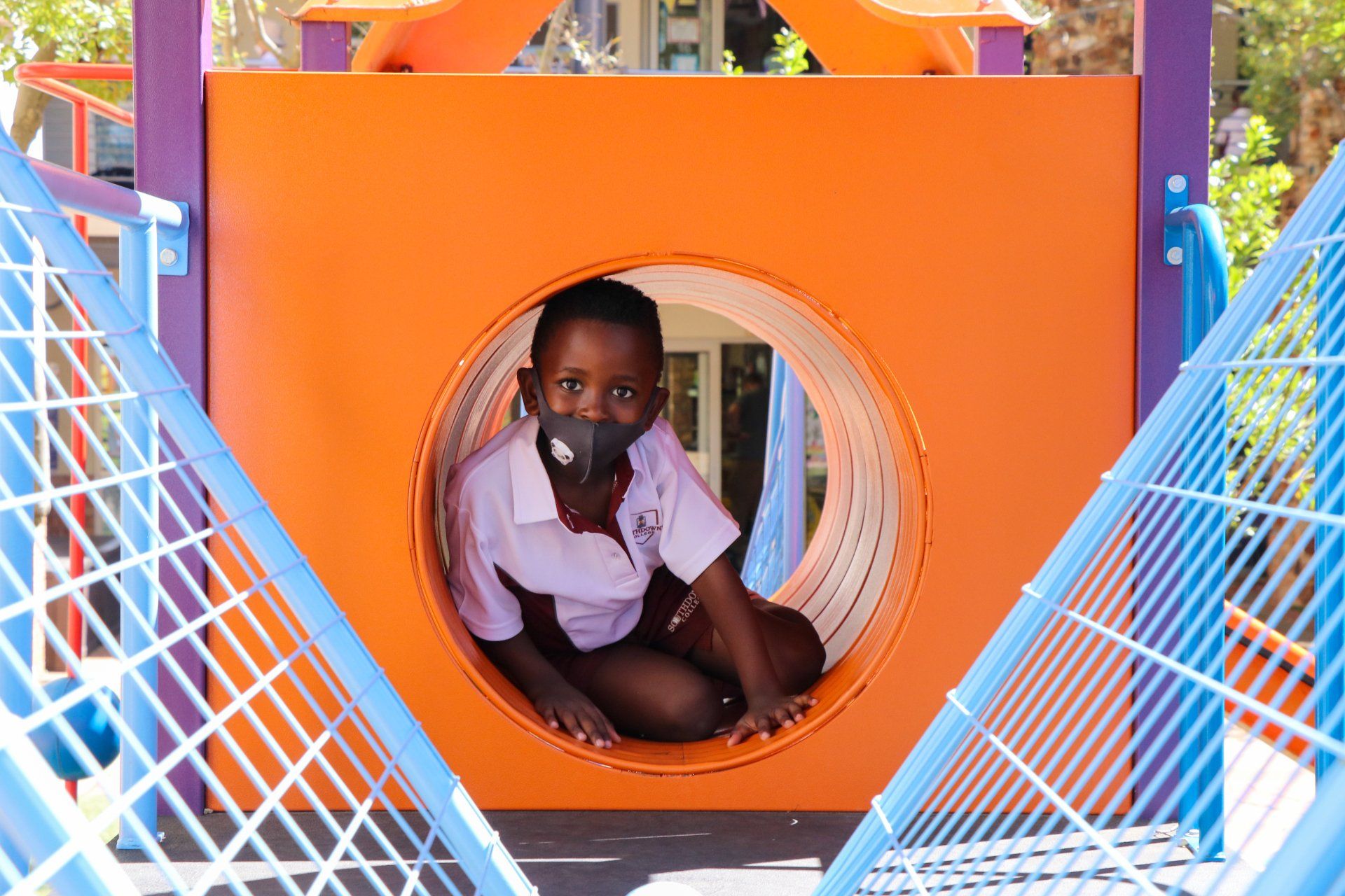 A young boy wearing a mask is sitting in a tunnel at a playground.