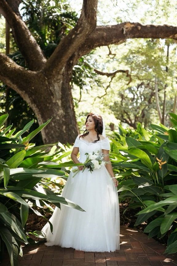 A Bride in a White Dress is Standing Under a Tree Holding a Bouquet of Flowers — Eternal Weddings & Couture In Woolner, NT