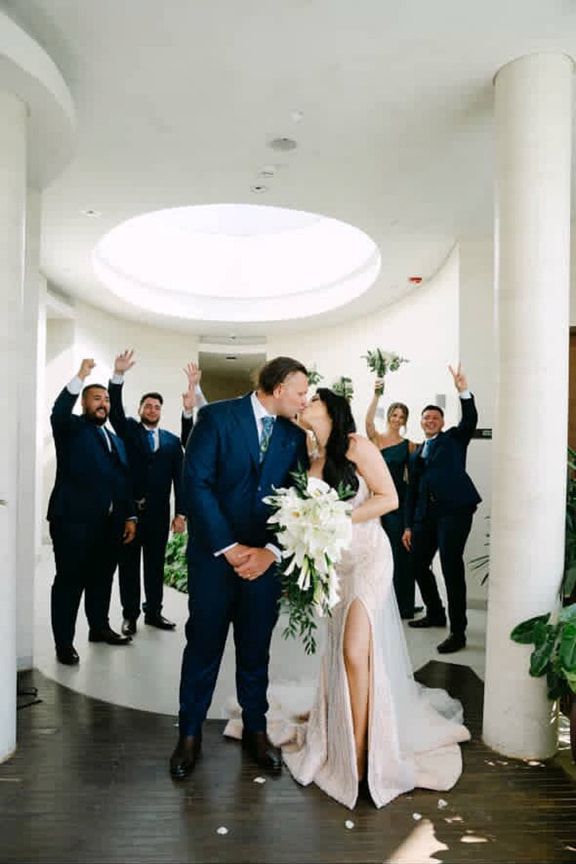 A Bride and Groom Kissing in Front of Their Wedding Party — Eternal Weddings & Couture In Woolner, NT