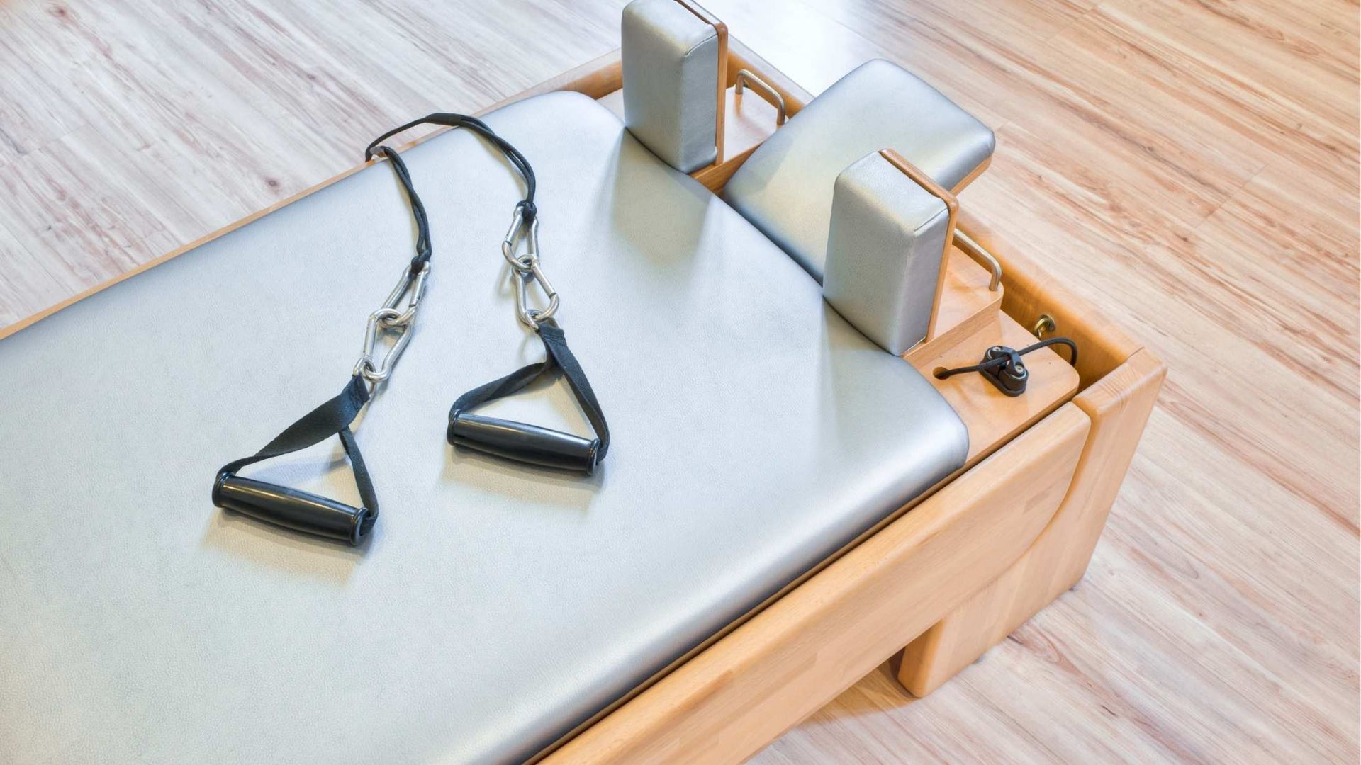 Pilates reformer bed with gray padded surface, straps, and headrest on a wooden frame.
