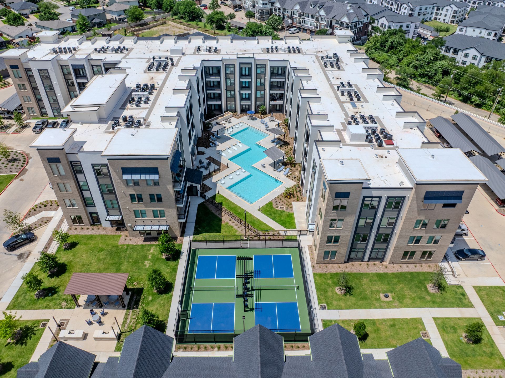 Aerial view of an apartment complex featuring a central courtyard with a swimming pool and a blue pickleball court.