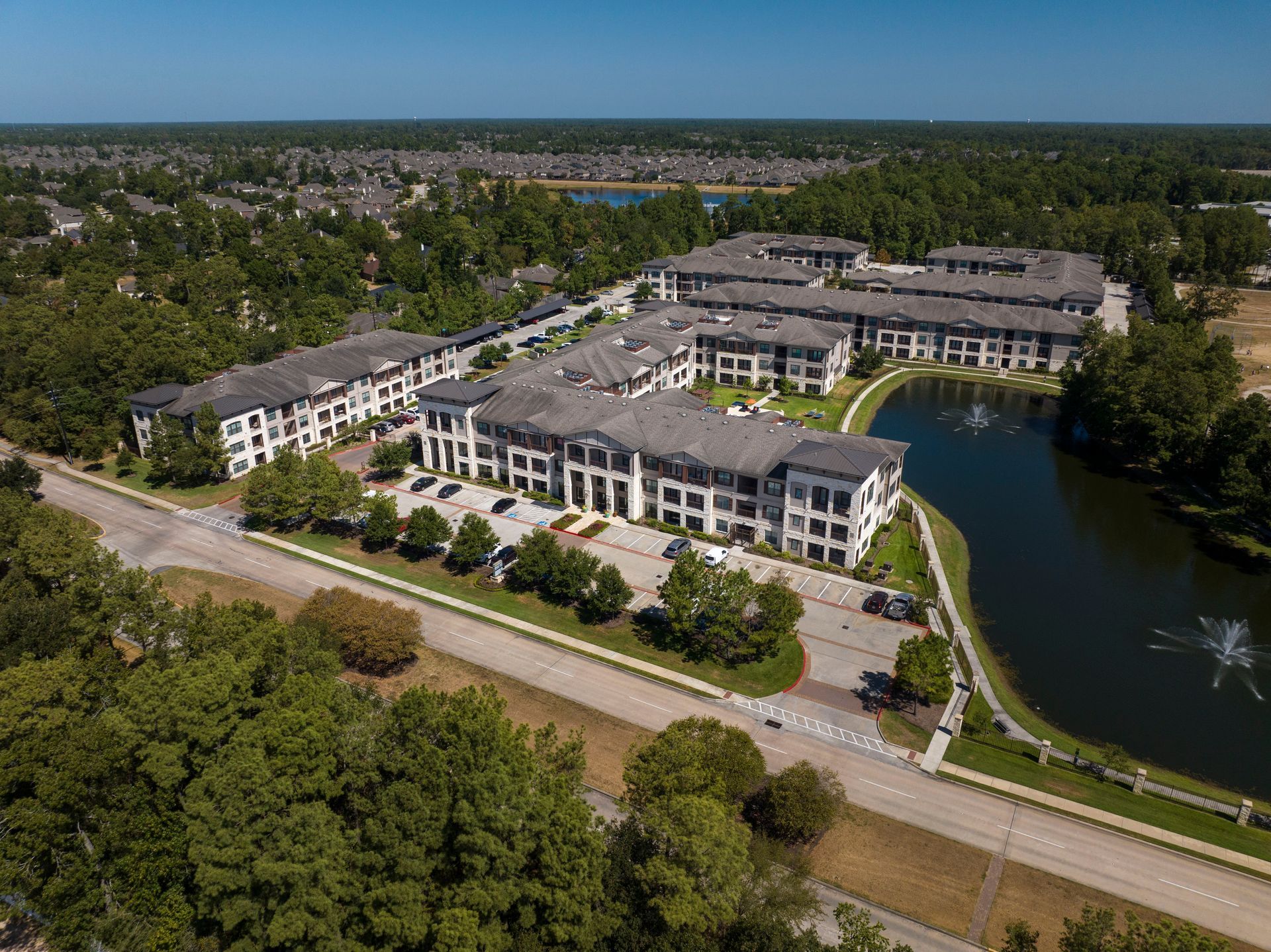 Aerial view of an apartment complex with multiple buildings, parking areas, and a small lake with fountains.