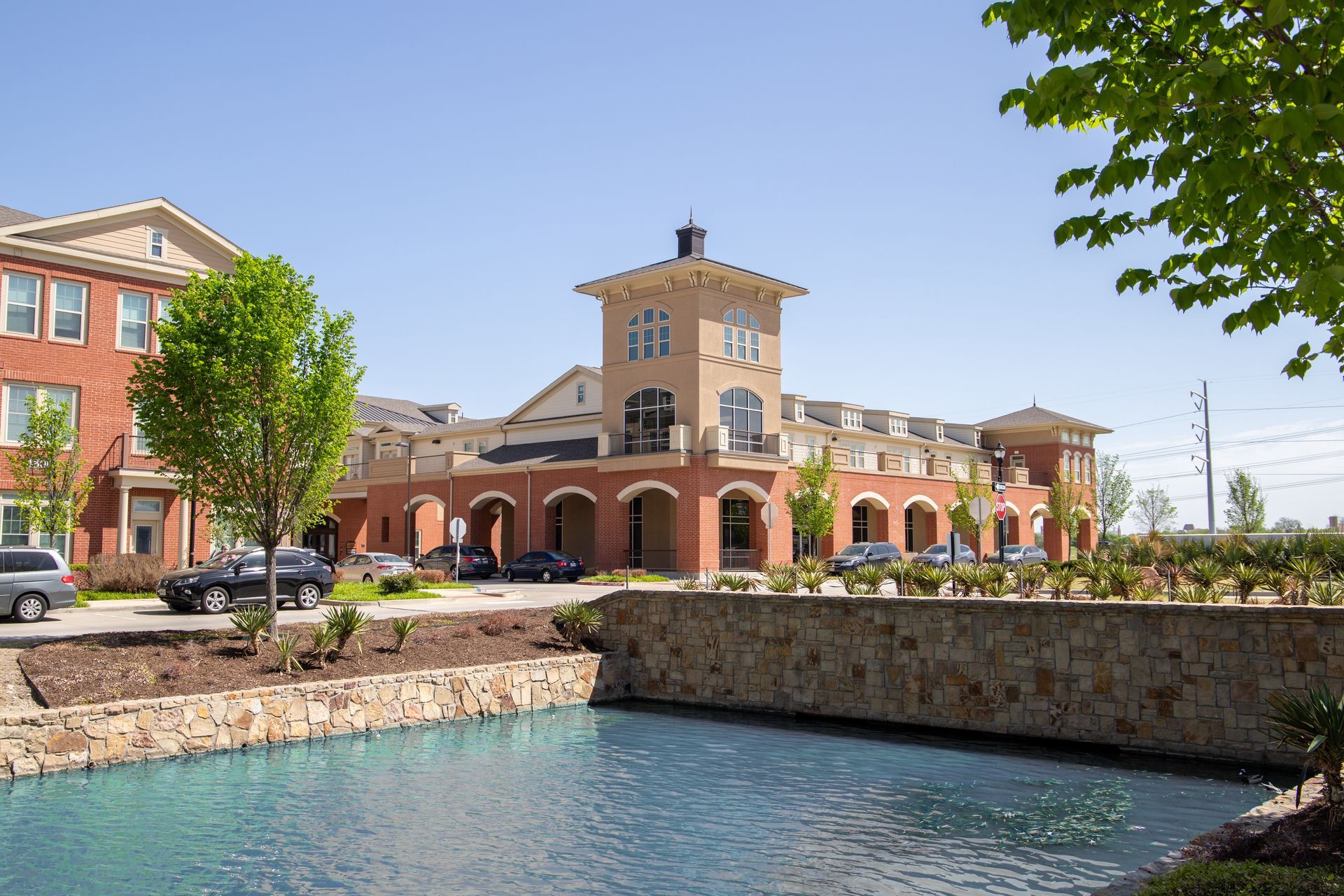 A modern brick building with a central tower stands behind a stone-walled pond on a sunny day.