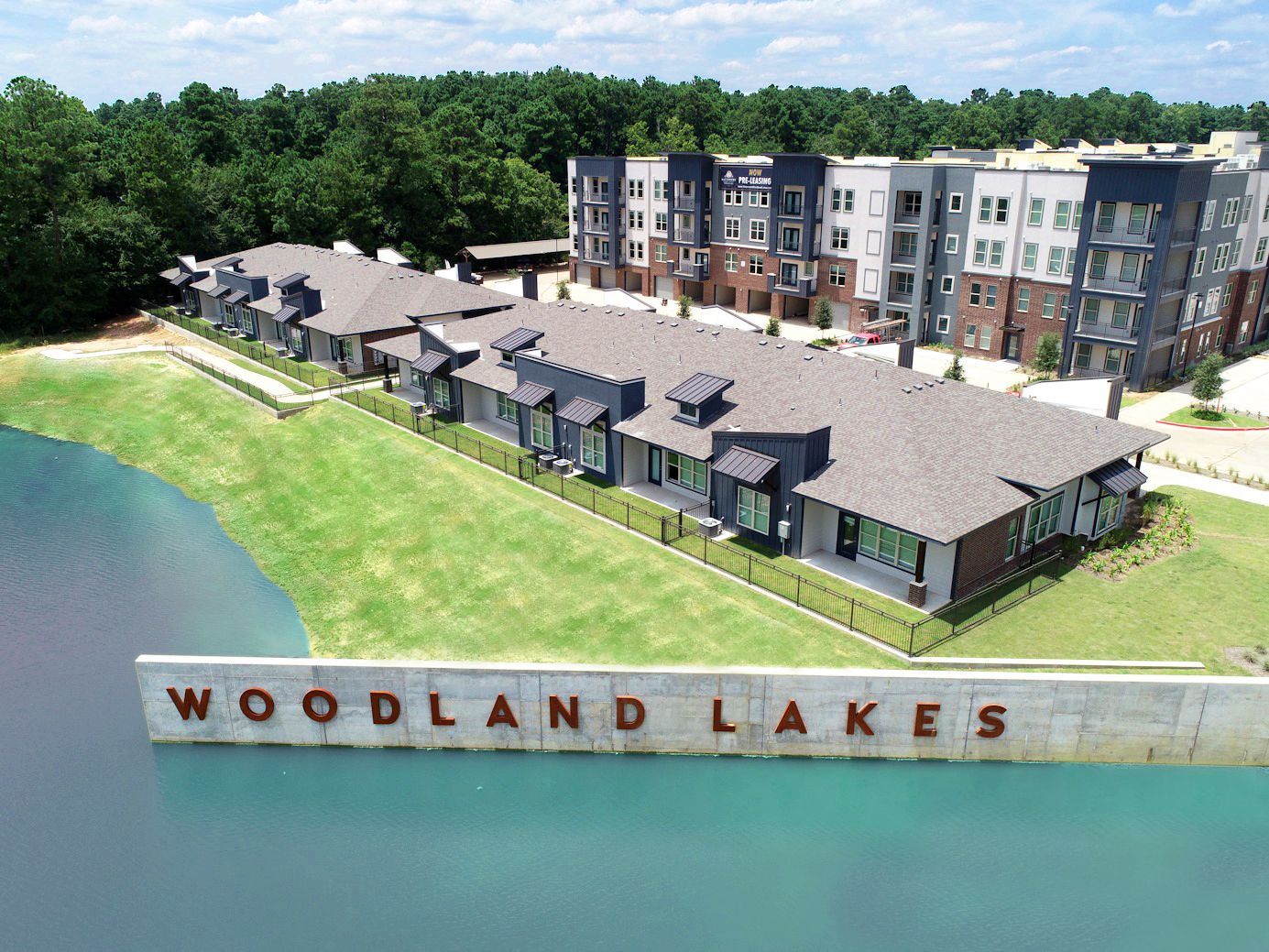 Aerial view of a Woodland Lakes apartment complex next to a lake with a concrete sign featuring the name in the water.