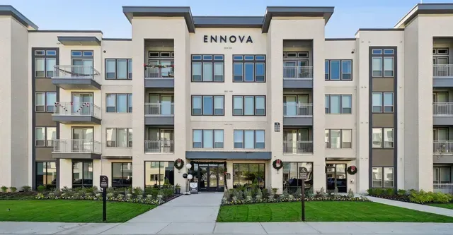 A four-story, modern apartment building with a light beige facade, gray accents, and green landscaping in front.