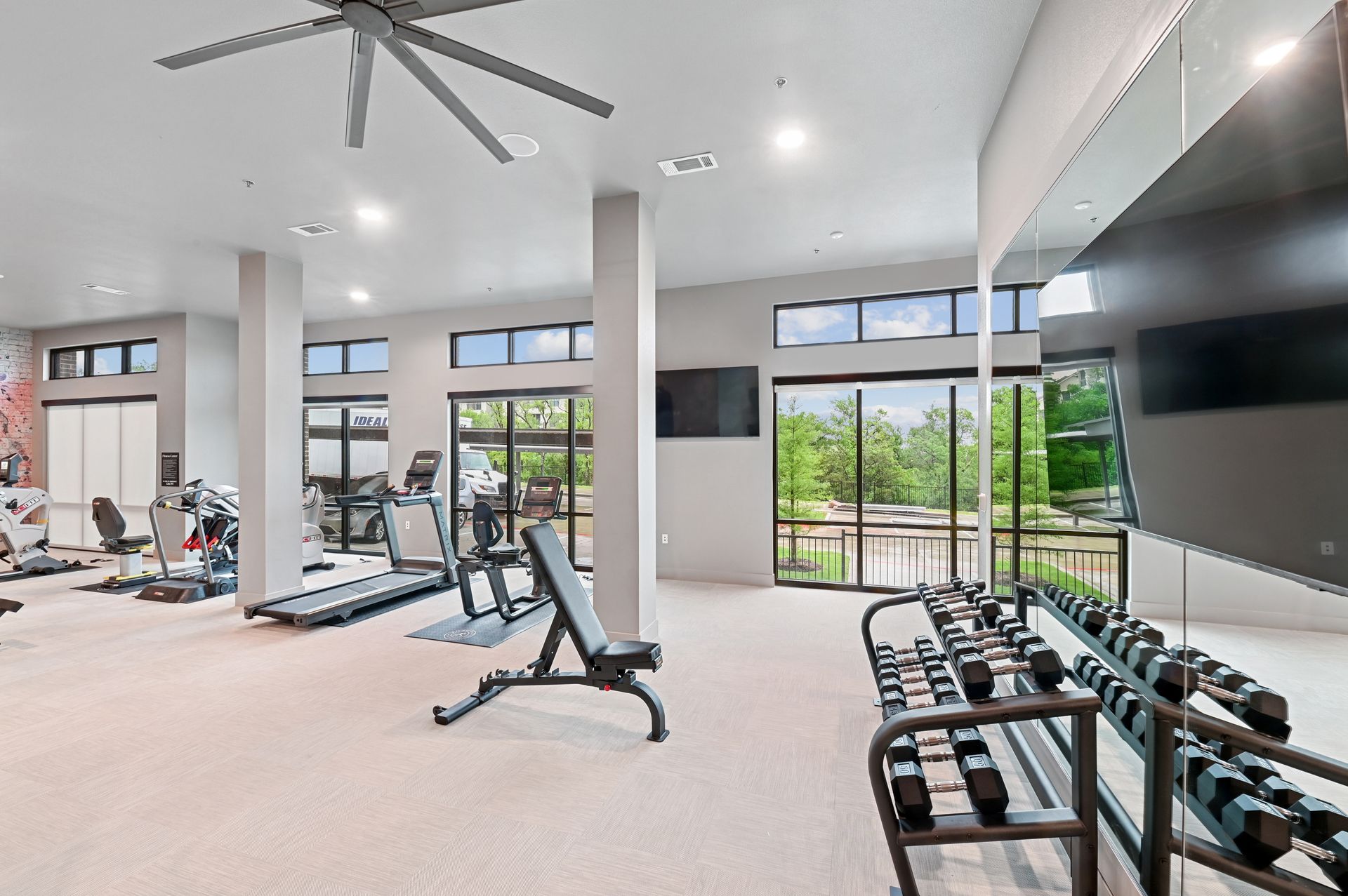 A brightly lit fitness center with gym equipment including treadmills, a bench, and a rack of dumbbells.
