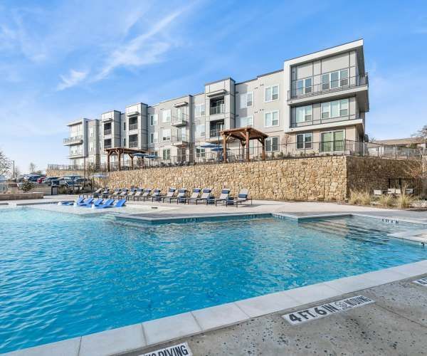 A bright blue swimming pool sits in front of a modern multi-story apartment building with a stone retaining wall.