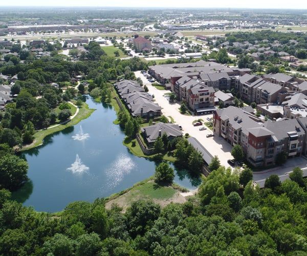 Aerial view of a residential apartment complex nestled next to a large pond with fountains, surrounded by lush green trees.
