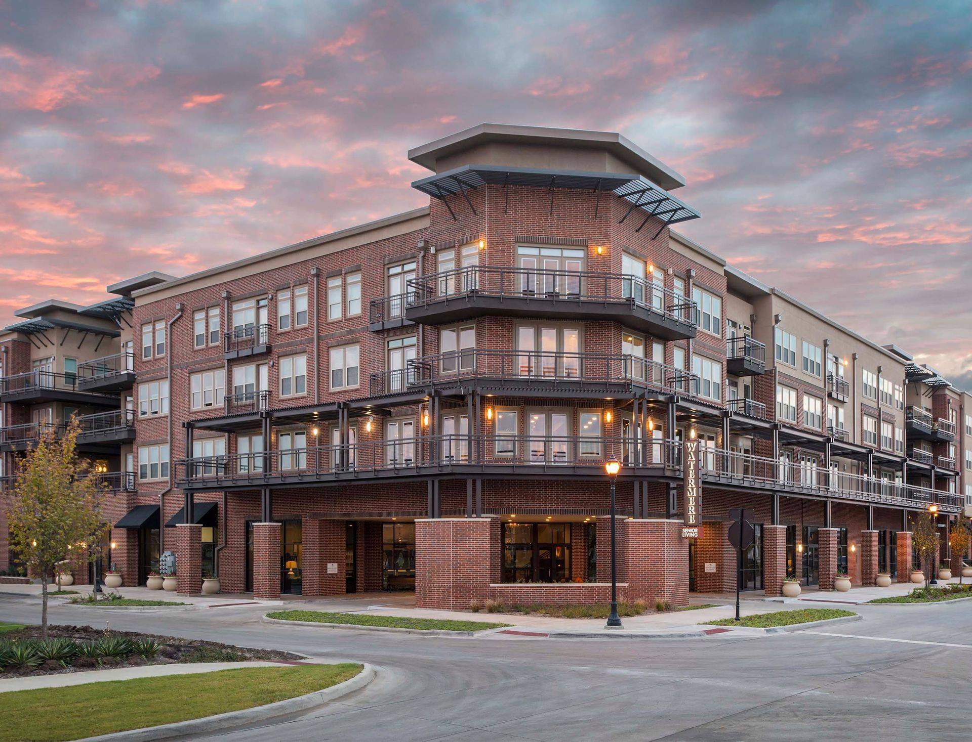 A multi-story brick apartment building with metal balconies under a cloudy pink and blue sunset sky.