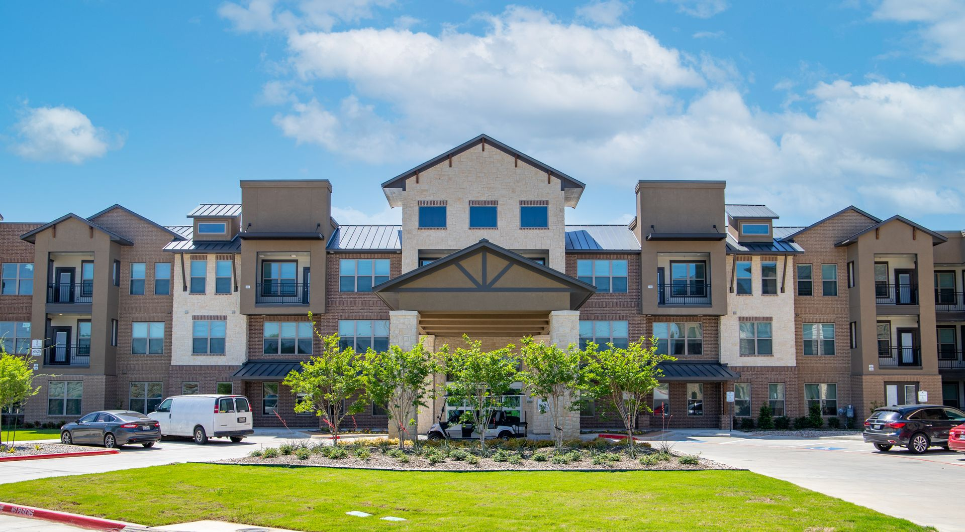 A three-story apartment building with a tan and brick exterior, a central entrance, and a green lawn under a sunny sky.