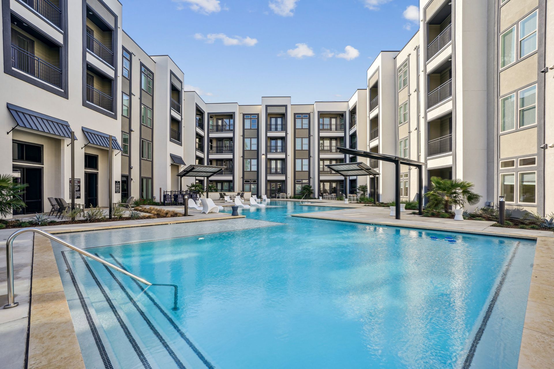 Modern apartment complex courtyard featuring a large swimming pool with steps, lounge chairs, and light-colored buildings.